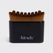 A Kit-Sch grooming brush with brown bristles and a black handle, featuring the Kit-Sch logo on the front, set against a plain light gray background.